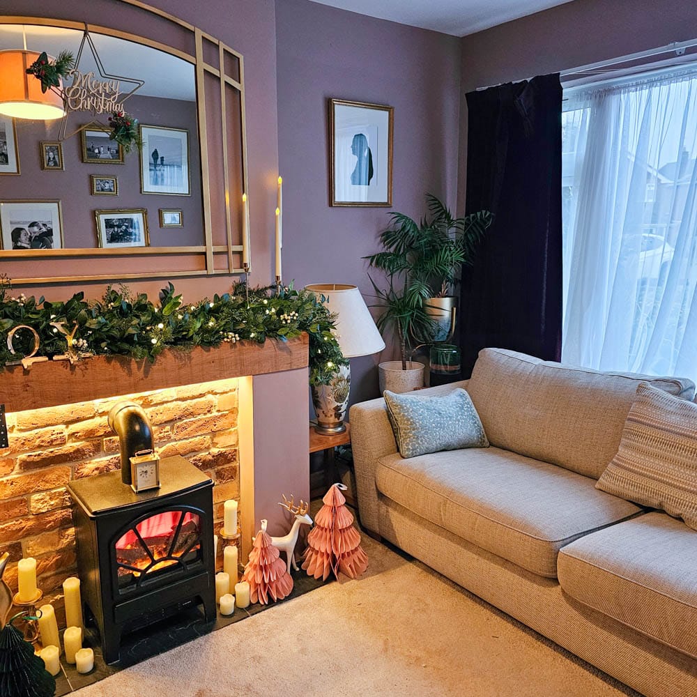 A cosy living room decorated for Christmas with garland and lights on the mantelpiece, candles and festive ornaments near a stove heater, a beige sofa, framed artwork on purple walls, and a Briiv Air Purifier adding fresh air—one year update review.