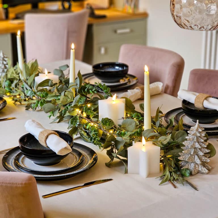 A festive dining table set with black plates, white serviettes, gold serviette rings, greenery garland, lit candles, and a glittery silver Christmas tree ornament creates stunning Christmas table décor. The background shows a cosy kitchen area.