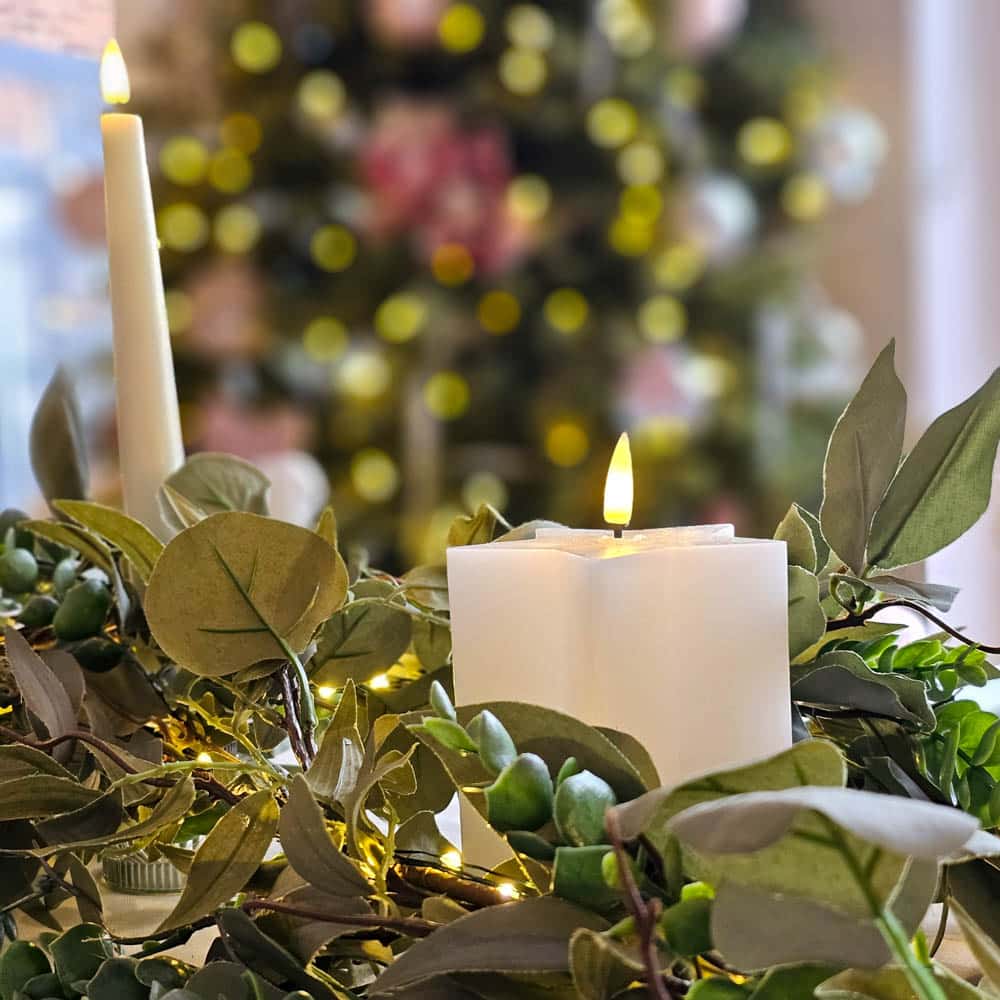 A close-up of leafy green foliage and two lit candles—one tall and one square—creates the perfect Christmas table décor against a blurred background of a decorated Christmas tree with twinkling lights.