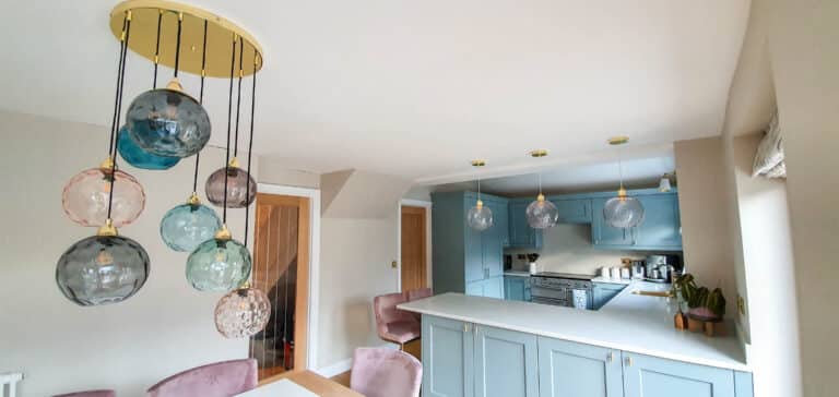 Modern kitchen and dining area with pastel blue cupboards, a white worktop, pink velvet chairs, and contemporary glass pendant lights—designed to keep your home warm while adding style and comfort.