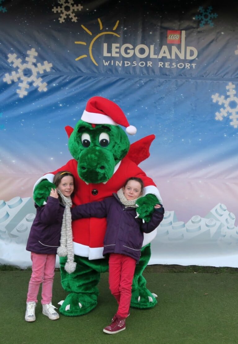 Two smiling children in winter clothes pose before a green dragon mascot in a Father Christmas outfit at LEGOLAND Windsor Resort, with snowy décor and sun—reminding visitors that maintaining furry inflatables like this mascot helps ensure holiday fun lasts all season.
