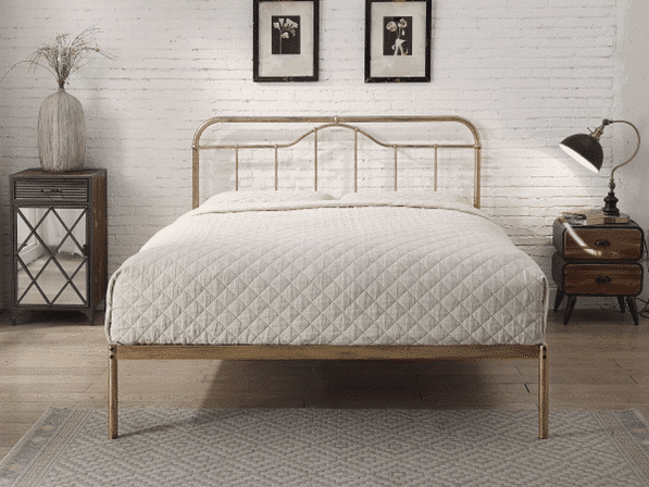 A neatly made bed with a light quilt, metal frame, and headboard stands between two bedside tables—one with a lamp and the other with a vase—showcasing mixed metals in your industrial bedroom. Two framed pictures hang on a white brick wall behind the bed.
