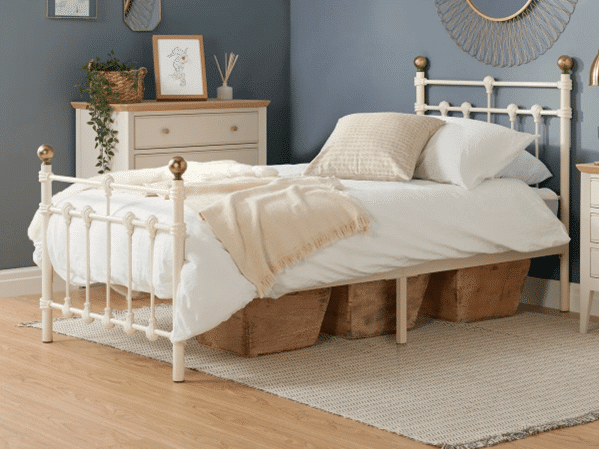 A neatly made bed with white and beige bedding and gold-topped posts stands on a woven rug, showcasing mixed metals in your industrial bedroom. Three wicker storage baskets are under the bed, with a chest of drawers, plant, and framed picture against a blue wall.