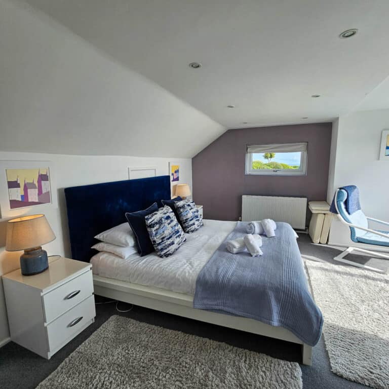 A cosy bedroom in family housing, featuring a double bed with a blue headboard, white bedding, and blue accents. Two bedside tables with lamps, a white desk, armchair, wall art, and a window overlooking greenery and blue sky.