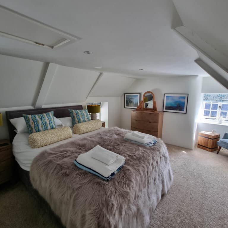 A cosy attic bedroom designed to maximise space, featuring a large bed with a fluffy grey blanket, colourful pillows, and folded towels. A wooden dresser with a mirror, framed art, and natural light make it feel just like home.