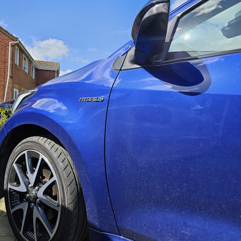 Close-up of a blue car’s front left side, showing the wheel, wing mirror, and a “HYBRID” badge powering the future. A red brick house and a blue sky with clouds appear in the background.