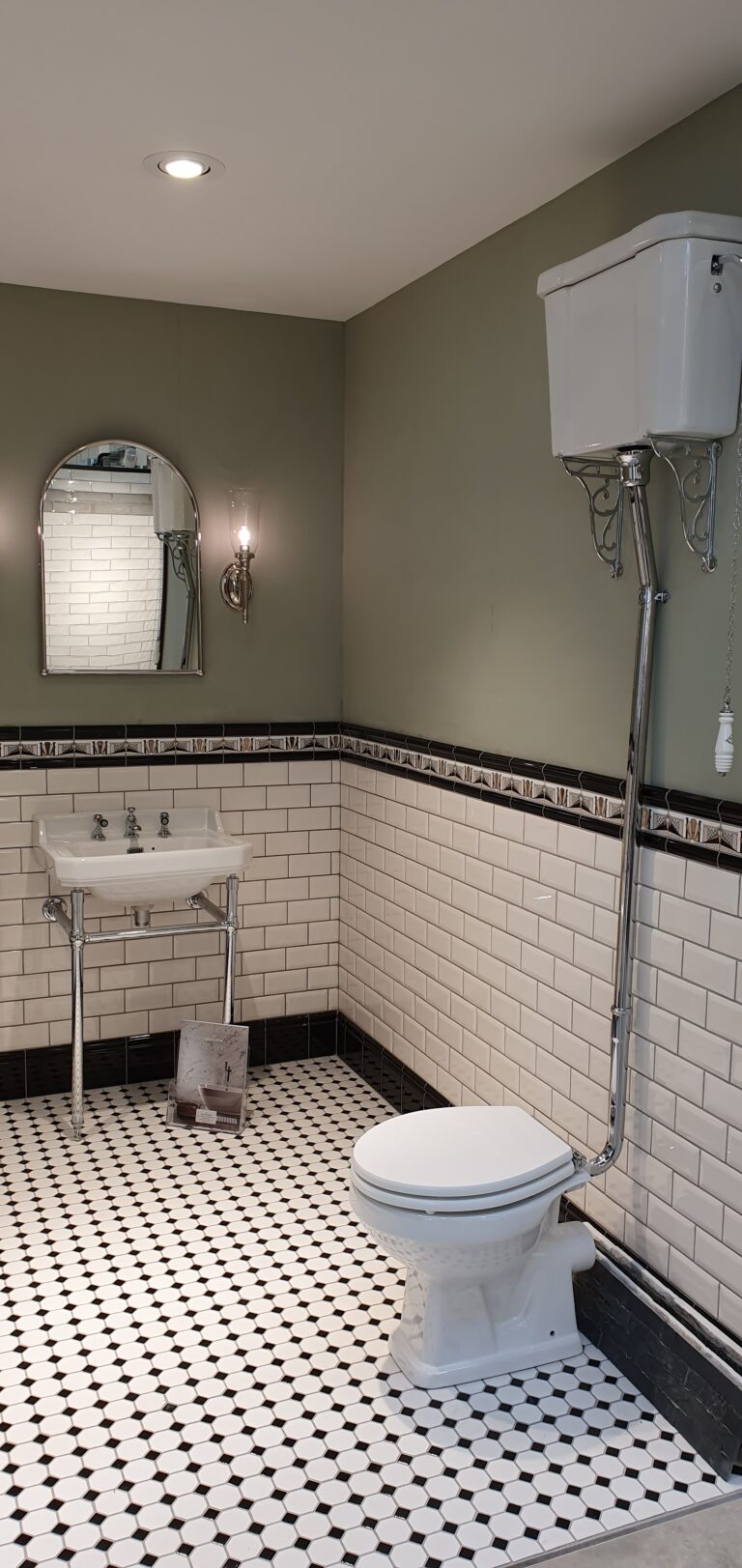 A vintage-style bathroom with white subway tile walls, black trim, and a pedestal basin features a luxurious design, highlighted by a high-level toilet with pull chain, mirror, wall sconce, and patterned black-and-white tiled floor.