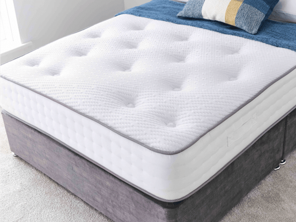 A neatly made bed with a white, tufted mattress—one of the best beds for summer—features a blue throw and a decorative cushion with white, blue, and yellow blocks in a bright, carpeted room.