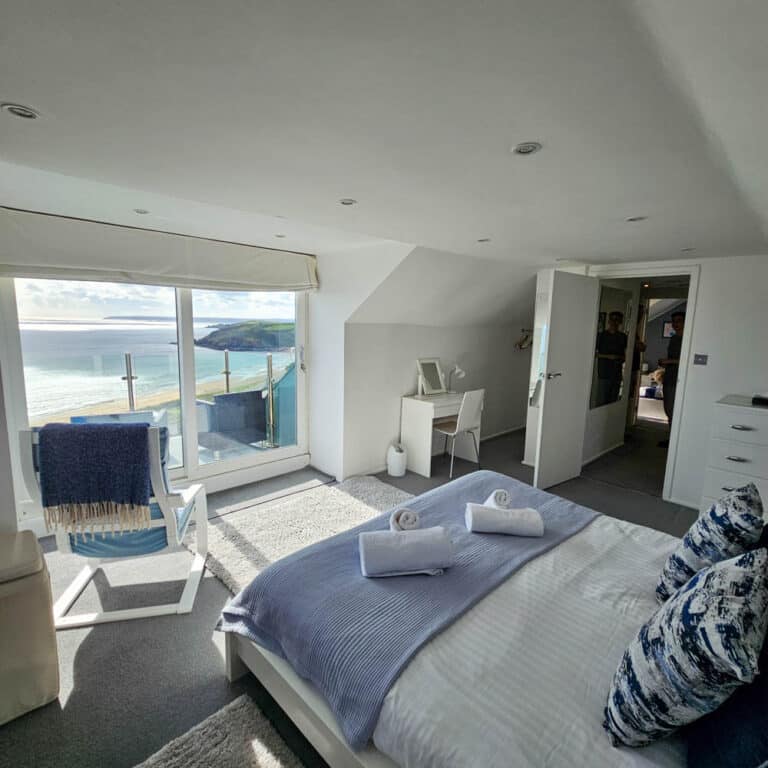 Bright bedroom with a double bed, blue and white bedding, and towels facing large windows overlooking a coastal view. A balcony with glass balustrade, chair, dressing table, and light decor create an airier space maximising light for a brighter home.