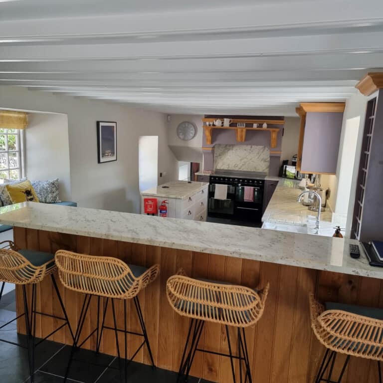 A modern kitchen with marble worktops, a wooden breakfast bar, four wicker barstools, fluted wall panels, a black cooker, and light cupboards. A cosy seating area with cushions is visible near a window in the background.