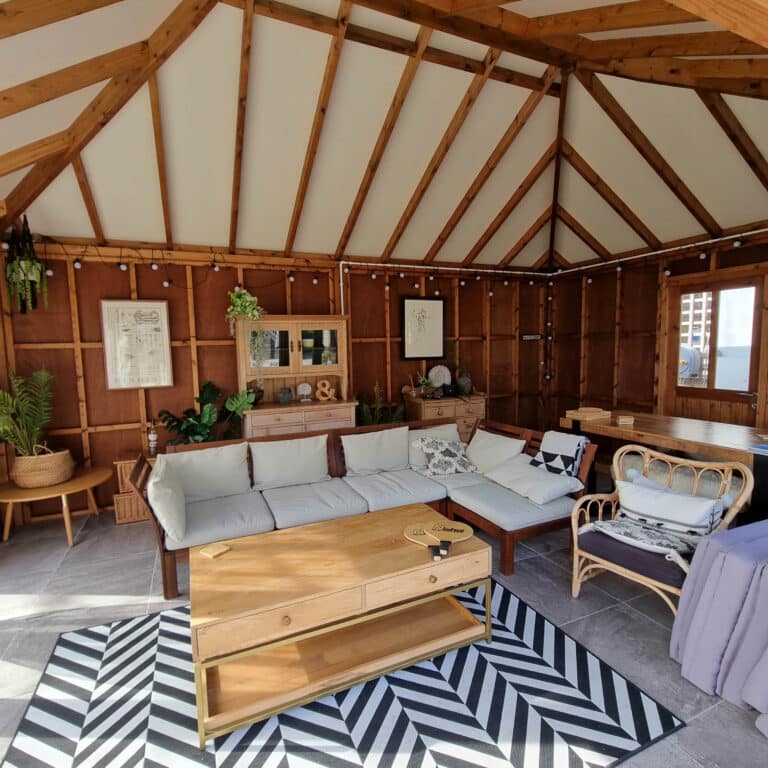 A cosy wooden room with exposed beams, featuring a white corner sofa of durable furniture, wooden coffee table, rattan chair, plants, fairy lights, framed art, and a black-and-white chevron rug on a tiled floor.