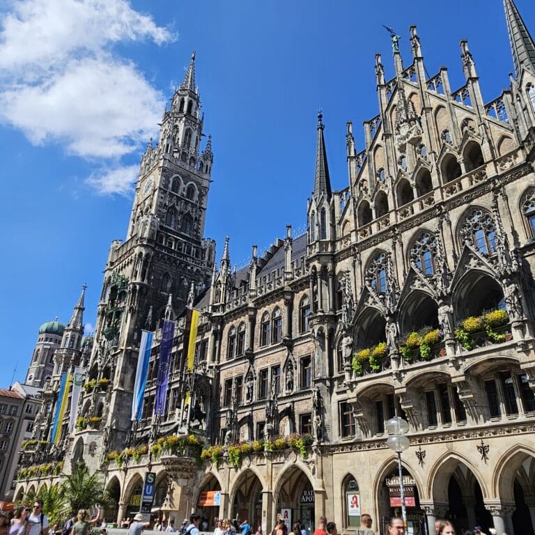 The image shows Munich’s New Town Hall with ornate Gothic architecture, flower boxes on windows, and colourful flags—highlighting a must-see for anyone looking to travel Europe on a budget whilst exploring lively Marienplatz under a bright blue sky.
