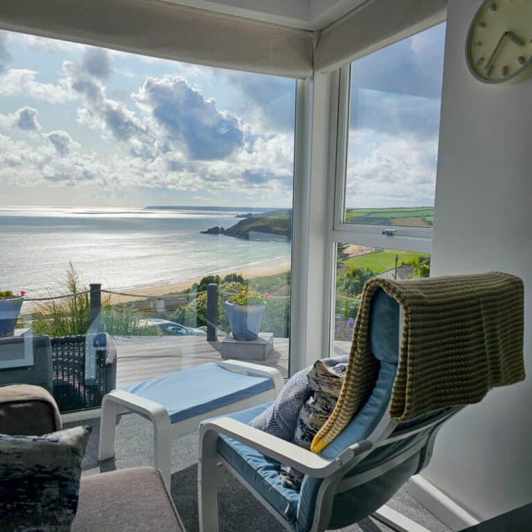 A cosy sunlit room with armchairs and a table overlooks a scenic beach and sea—essential criteria for any property search—with green hills, a partly cloudy sky beyond large windows, and a clock on the white wall to the right.