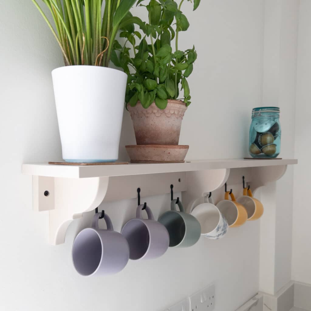 A wall-mounted shelf adds kitchen functionality, holding potted plants and a jar of stones on top, with five colourful mugs hanging from hooks underneath. The shelf brightens up the white-walled room, reflecting modern kitchen interior design.