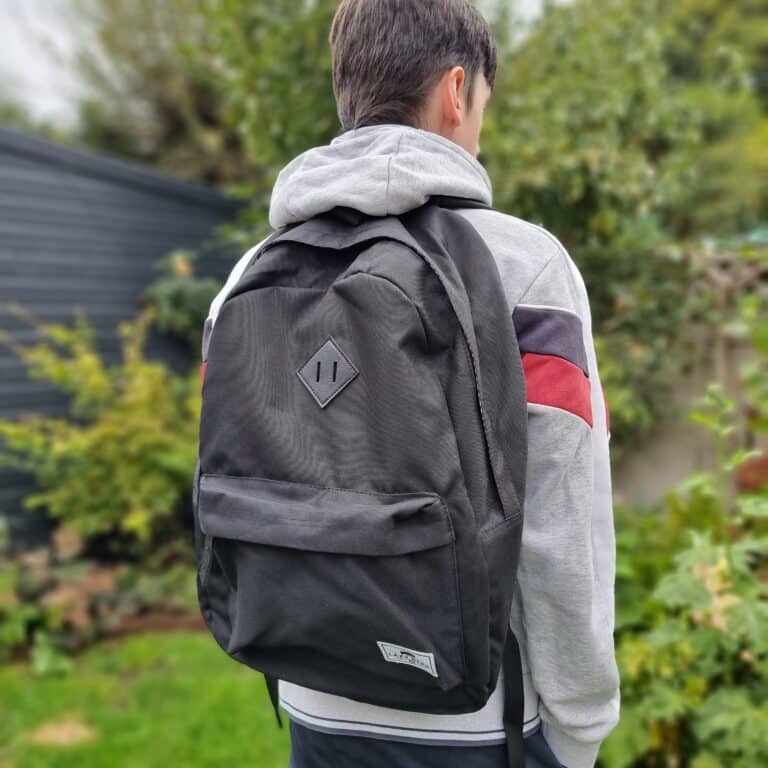 A person wearing a grey hoodie with red and navy stripes stands outdoors, carrying a large black rucksack—perhaps filled with coursework or A-Level revision tips. Green plants and a dark fence are visible in the background.