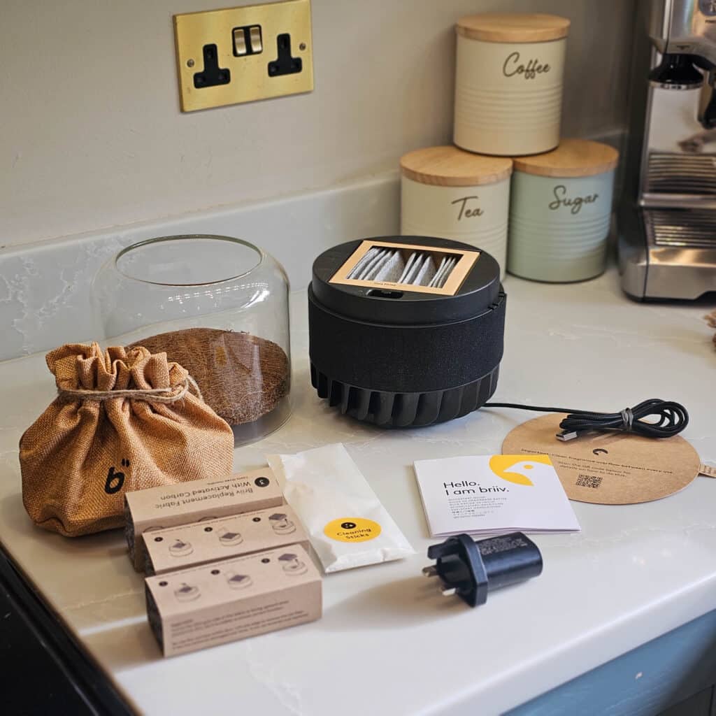 A worktop with a coffee maker, labelled jars for coffee, tea, and sugar, and a modern black appliance—possibly a sustainable air purifier like the Briiv 2 Pro—surrounded by accessories including a plug adaptor, cables, boxes, burlap pouch, and manuals.