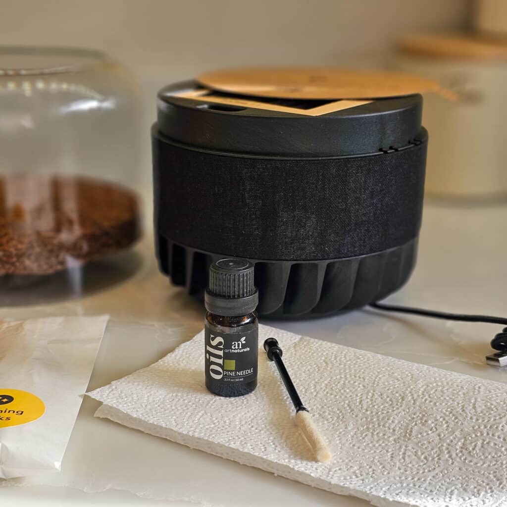 A small essential oil bottle, a dropper, and a paper towel sit on a worktop near a black diffuser, a glass jar of coffee grounds, and the Briiv 2 Pro—a sustainable air purifier often featured in top air purifier reviews.