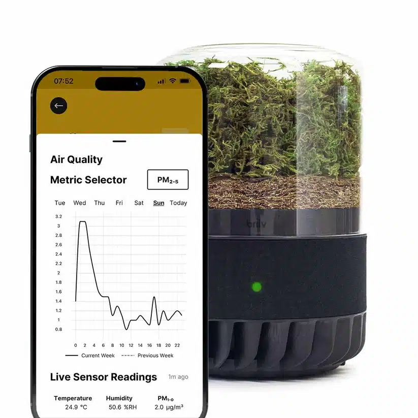 A smartphone displays air quality metrics and sensor readings next to the Briiv 2 Pro, a modern air purifier featuring visible green moss inside a clear container. The purifier has a round black base with a glowing green indicator light.