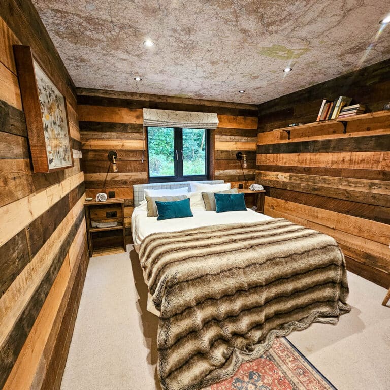 A cosy log cabin bedroom with wood-panelled walls, a double bed draped in a fur blanket and teal pillows, bedside tables with lamps, a window framing greenery, and shelves featuring books and decor.