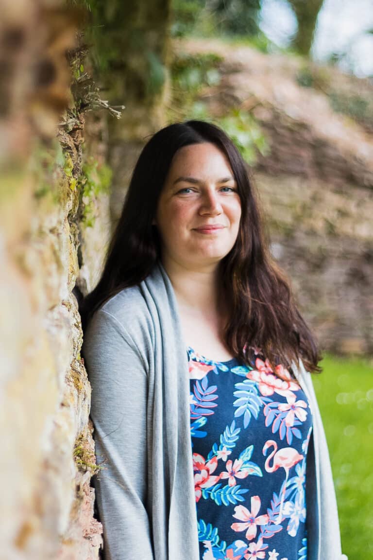 A woman with long dark hair and a natural look, wearing a grey cardigan and floral dress, stands outside leaning against a stone wall, smiling softly. The photos capture her against blurred greenery.