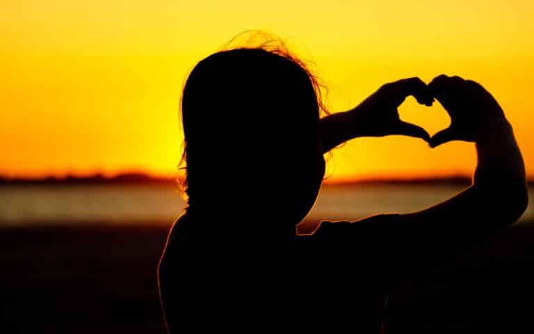 A silhouette of a person at sunset, holding their hands up to form a heart shape against the bright orange and yellow sky near a body of water, captures the serene beauty often found near hidden culinary gems or charming restaurants.