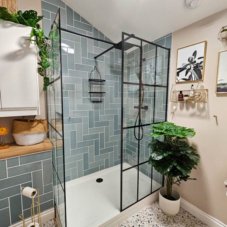 Modern bathroom with a glass shower enclosure, black metal frame, and blue herringbone tiles. Stylish bathroom decor includes green plants—plus artificial plants for easy upkeep—wall shelves with decor, and a white cupboard with baskets and towels.