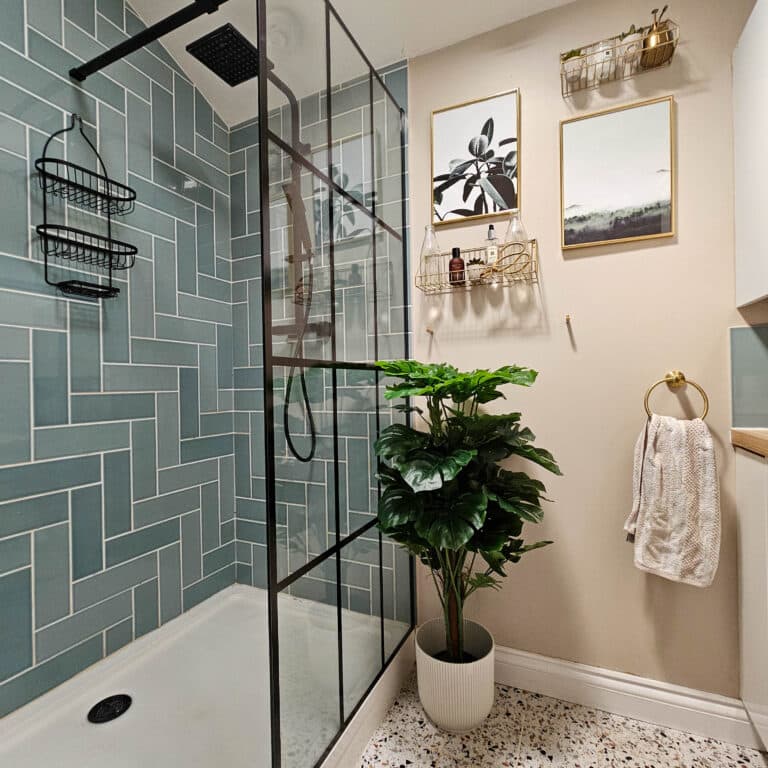 A modern bathroom with teal herringbone shower tiles, black fittings, a potted artificial plant, stylish wall shelves for bathroom décor and toiletries, a towel ring with a white towel, and terrazzo flooring—a perfect home improvement touch.