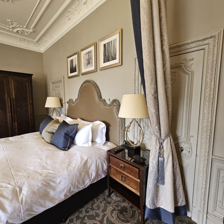 Elegant bedroom with a large, tufted headboard—perfect inspiration if you’re consulting a four-poster bed guide—to bedside tables with lamps, ornate moulding, framed art above the bed, and heavy curtains to the right.