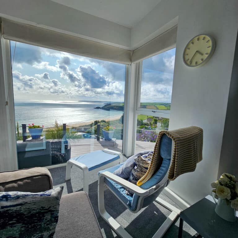 A cosy sunlit room with large modern windows overlooks the sea and coastal landscape. A chair with a blanket and cushion faces the view; a clock, potted plants, and flowers refresh home with a calming, relaxing atmosphere.
