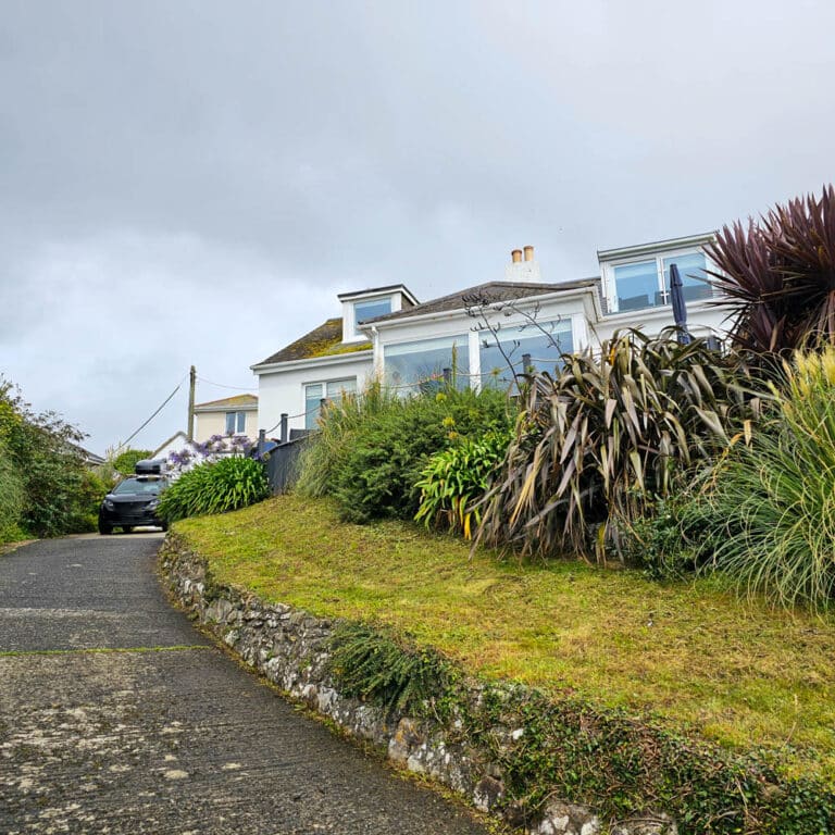 A sloped driveway leads up to a white house with large windows and modern cladding, surrounded by a garden full of shrubs and tall plants. A black car is parked near the home owner’s residence under an overcast sky.