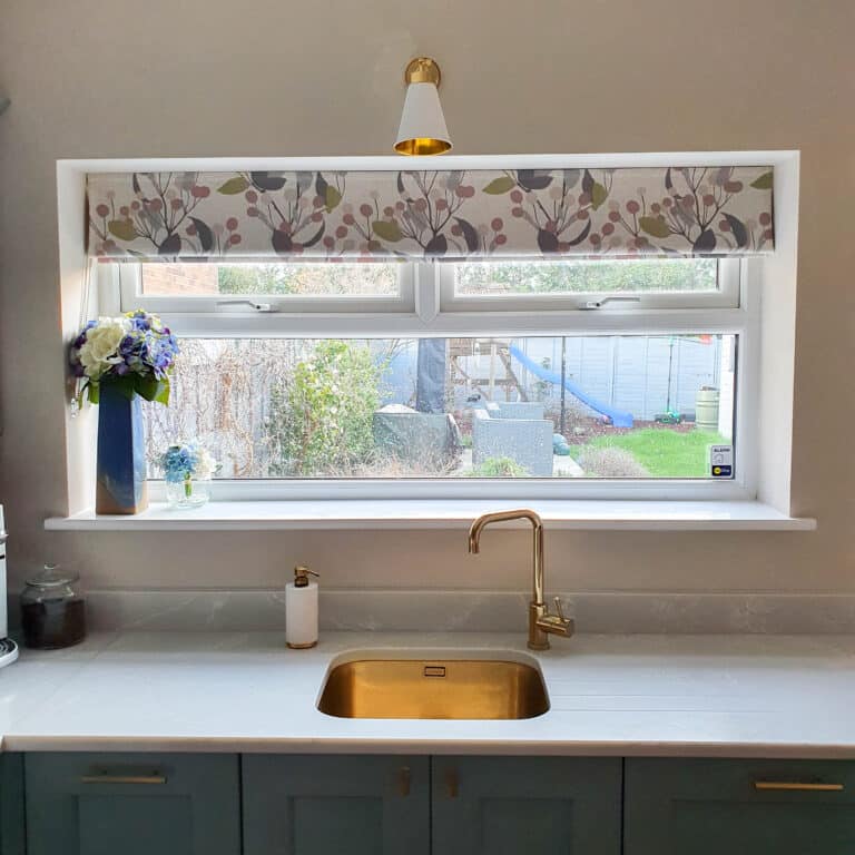 A tidy kitchen sink area with a gold tap and sink, a roll of kitchen paper, and a wide window framed by a floral valance. A vase of flowers sits on the sill, while uPVC doors in the North East ensure durability and style for your home.