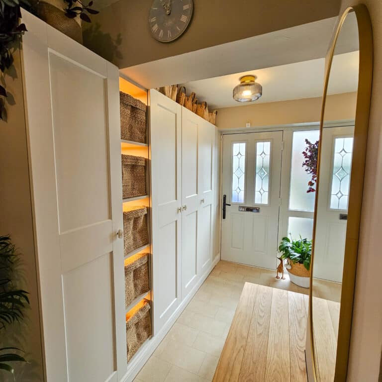 A bright entrance hall with white cupboards, an IKEA Pax hallway storage cupboard, shelves with woven baskets, a wooden floor, a round mirror, and a front door with frosted glass panels. A potted plant sits by the door beneath a wall clock.
