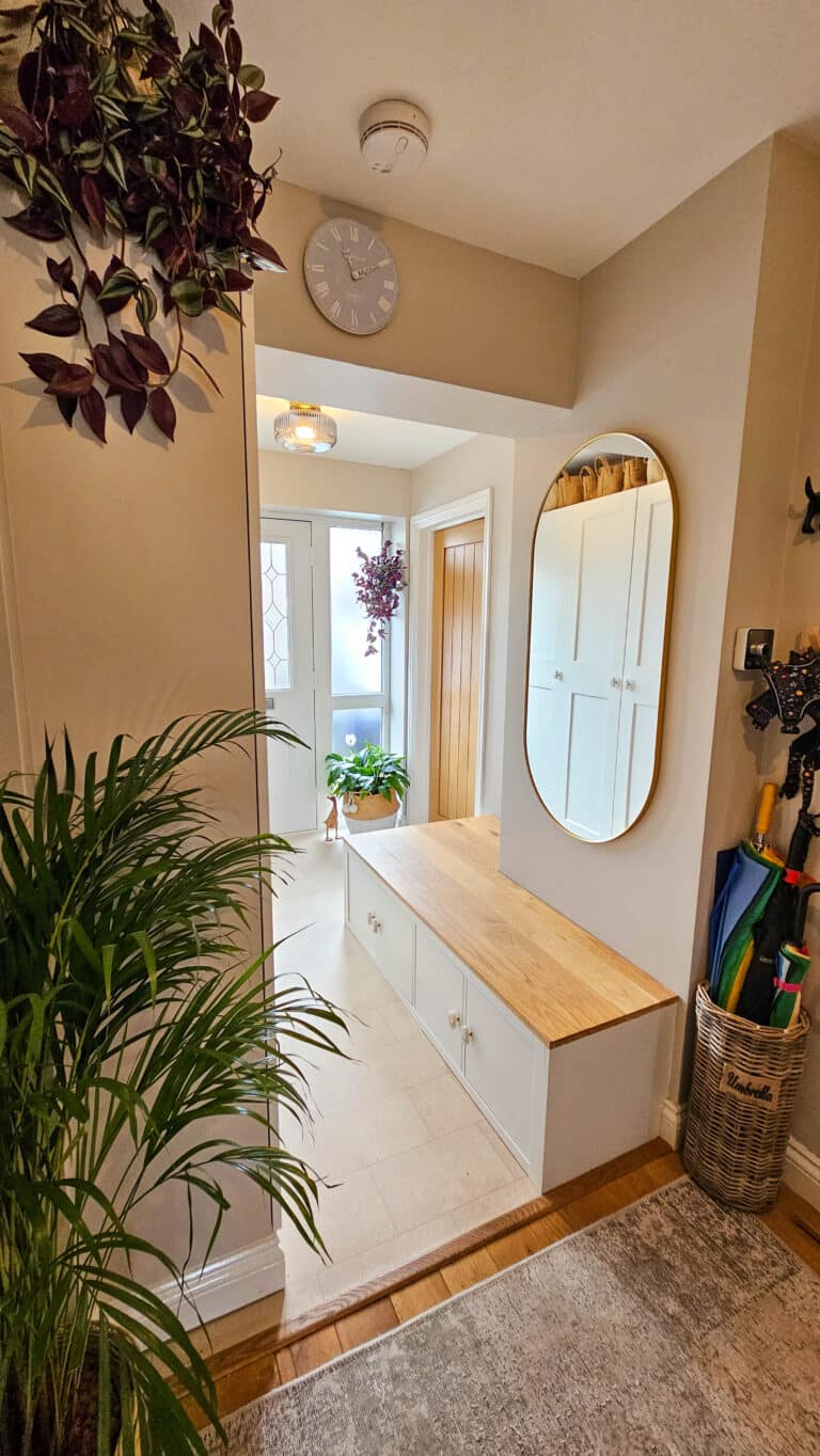 A bright, tidy entrance with white cupboards, IKEA Pax hallway storage, a wooden bench with storage, a round wall mirror, plants, umbrellas in a basket, and a wall clock. A glass door lets in natural light and displays a hanging plant outside.