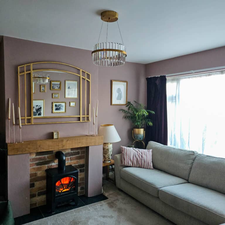 A cosy living room with a grey sofa, a lit wood-burning stove under a wooden mantelpiece, framed photos and art on mauve walls, a Pagazzi Camburn brushed gold LED pendant chandelier, a potted plant, and sunlight streaming through sheer curtains.