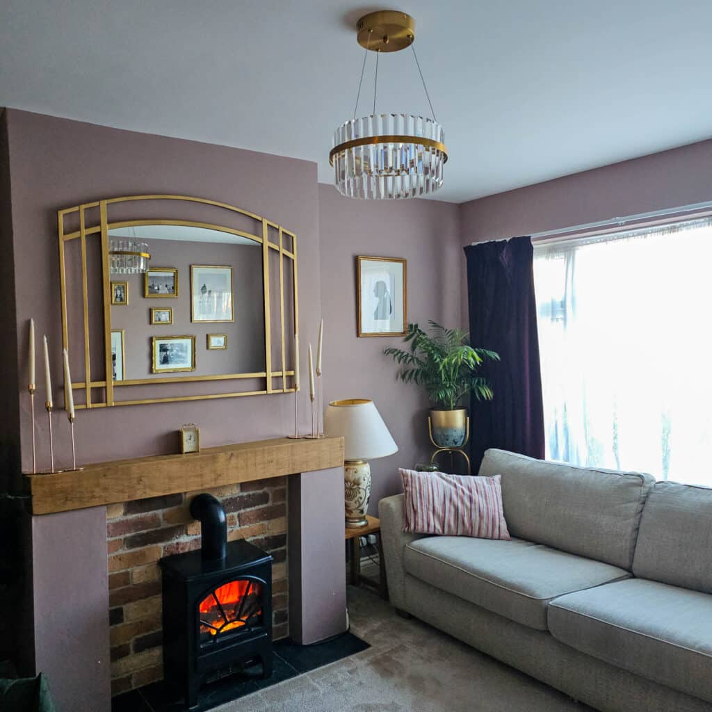 A cosy living room with mauve walls, a beige sofa, striped cushion, large window with sheer curtains, and a Pagazzi Camburn brushed gold pendant adding elegance above the wooden mantelpiece, framed artwork, potted plant, table lamp, and black electric fireplace.