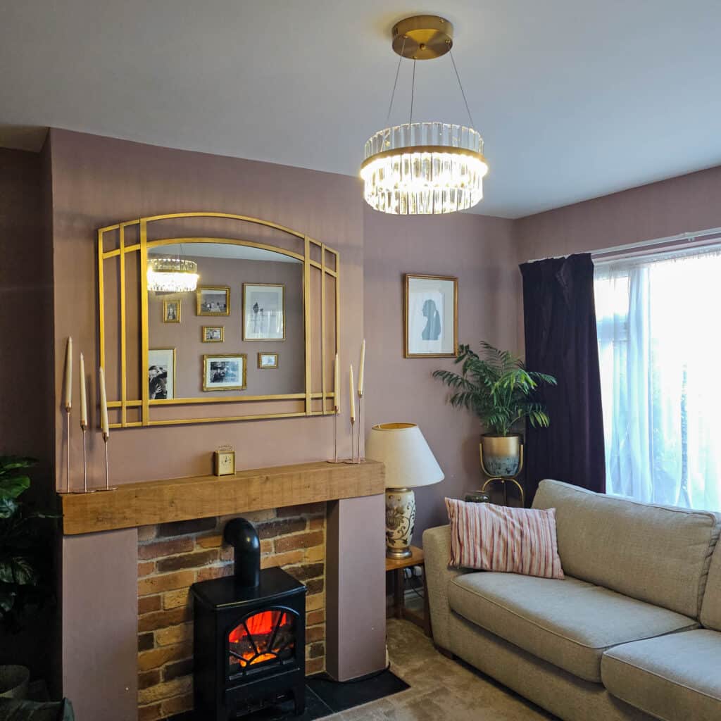 A cosy living room features a brick fireplace with a wooden mantel, a decorative mirror, framed art, a potted plant, and a beige sofa with a striped cushion, all illuminated by natural light and a Brushed Gold Halo LED Pendant.