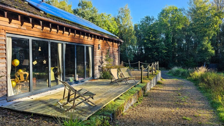 A wooden cabin with large glass doors opens onto a sunlit deck with loungers, surrounded by greenery and trees, under a clear blue sky—the perfect retreat for long-term roof care and saving tiles.