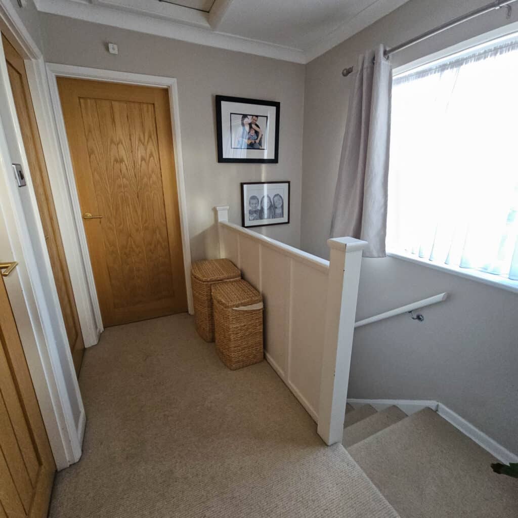 A carpeted landing at the top of a staircase with two wooden doors, a family laundry basket, wicker baskets, framed pictures on the wall, and a window with white curtains letting in natural light.