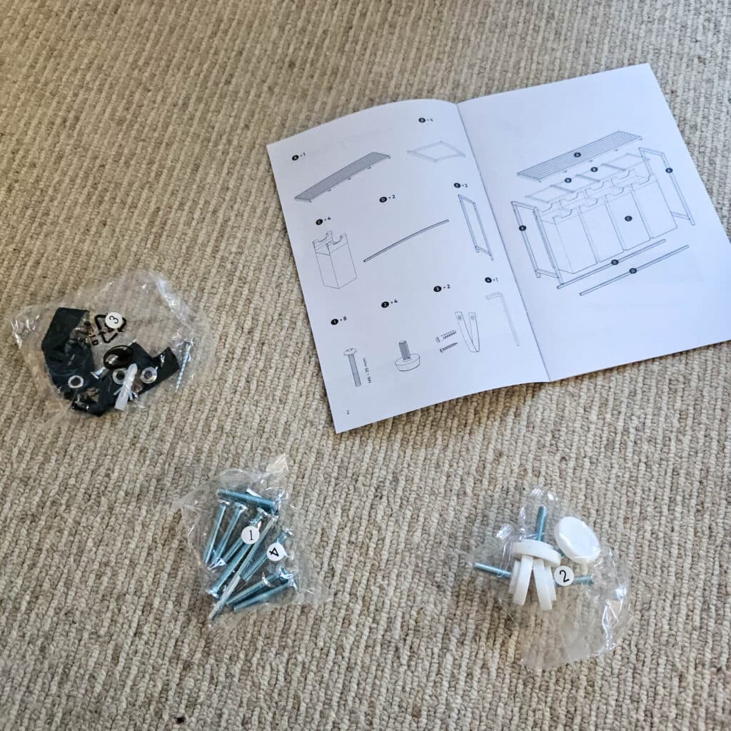 Three bags of screws and hardware are laid out on a beige carpet next to an open instruction manual, ready for assembling your VASAGLE laundry basket.