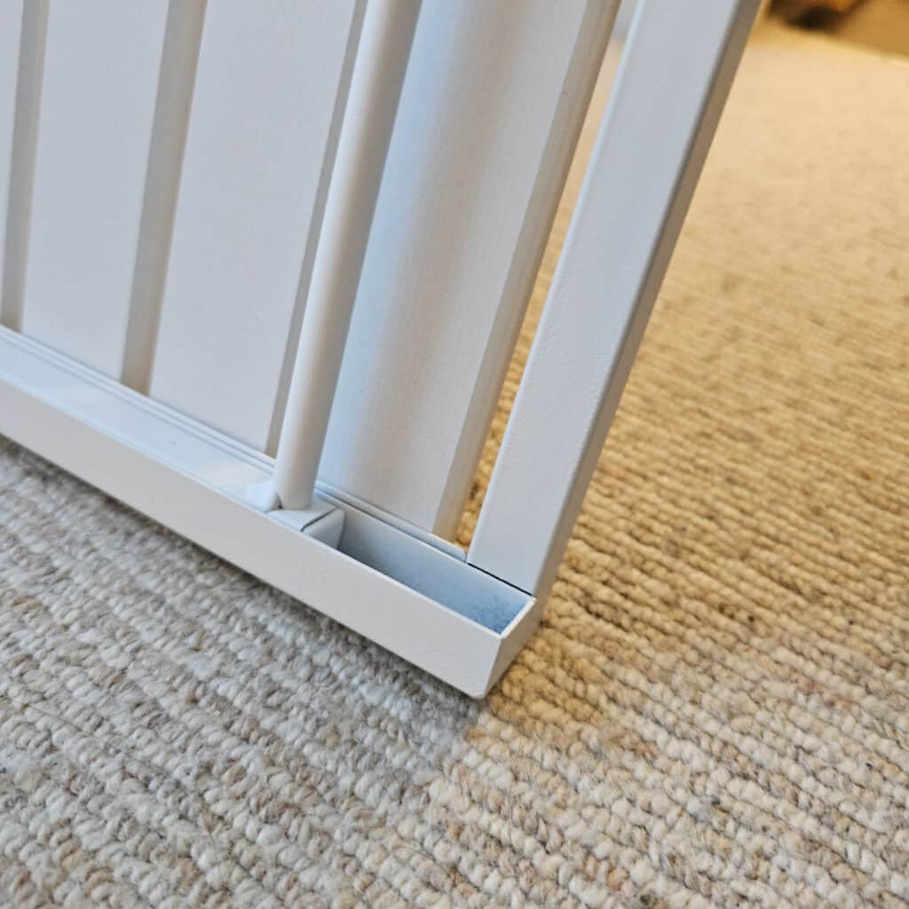 Close-up of the bottom corner of a white metal gate resting on a beige, textured carpet. The image highlights the gate’s bars and base detail, complementing a home organised with the Best Laundry Basket.