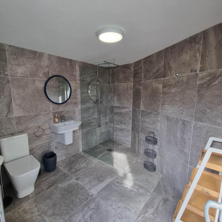 Modern bathroom with large grey tiles, a wall-mounted basin with a round mirror above, a glass walk-in shower, and natural light—this stylish living space promotes sustainable living with a metal shelf and wooden towel rail.