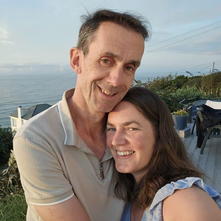 A smiling man and woman embrace outdoors on a sunny day, with a house, lush greenery, and the sea in the background—showcasing the joy of a healthy smile thanks to orthodontics.
