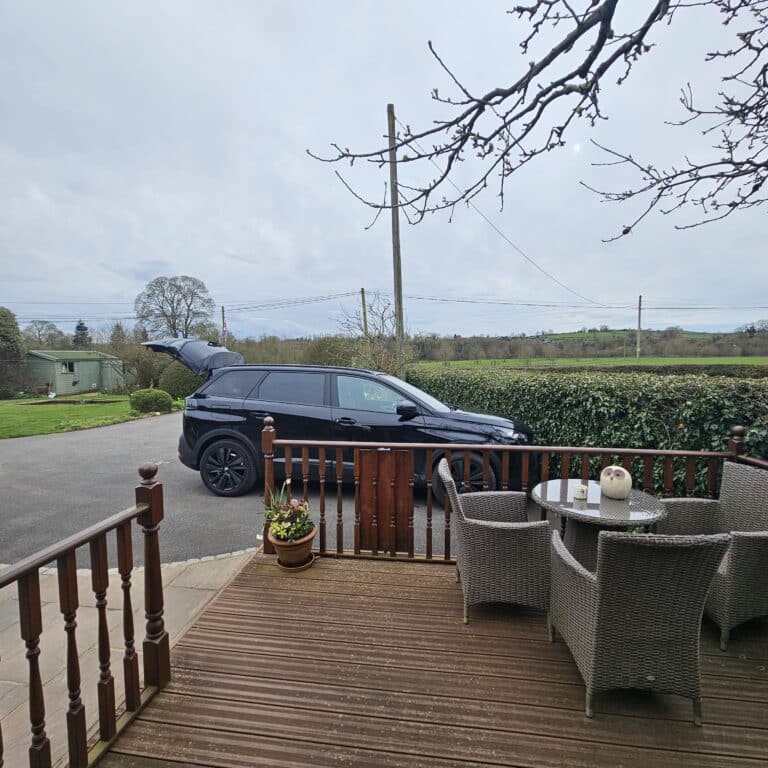 A black SUV with its boot open is parked on a driveway near a wooden decking with wicker chairs and a glass table—an inviting spot for smart travel planning to help save on travel whilst enjoying scenic views of trees, fields, and a cloudy sky.
