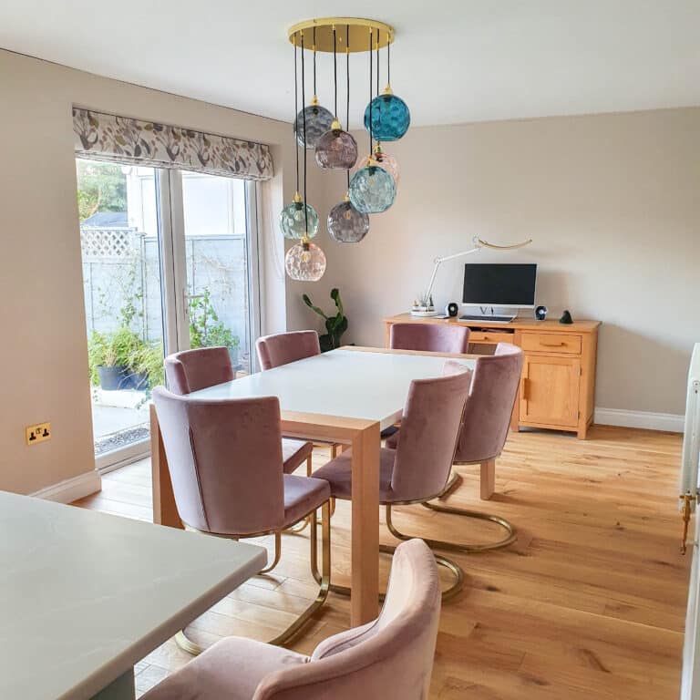 A modern dining room with a light wood floor—showcasing the advantages of Click Luxury Vinyl—pale walls, a white table, mauve velvet chairs, colourful glass pendants, a sideboard, and a computer on a desk near windows with floral blinds.