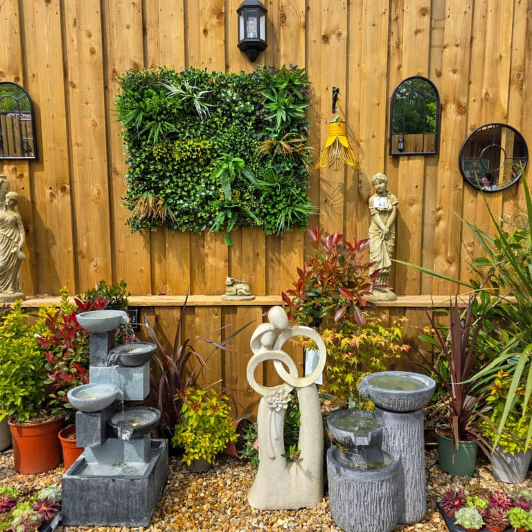 A garden display at Dobbies Cadnam features stone fountains, a modern abstract sculpture, and potted plants in front of a wooden fence updated after refurbishment. The fence has mirrors, statues, a lantern, and a lush green vertical wall planter.