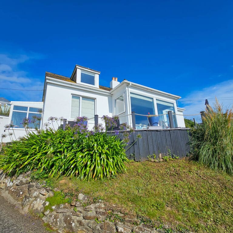 A white house with large windows and a sloping roof sits on a grassy hill, surrounded by green plants and flowers under a bright blue sky—showcasing the benefits of regular roof cleaning for any UK householder.