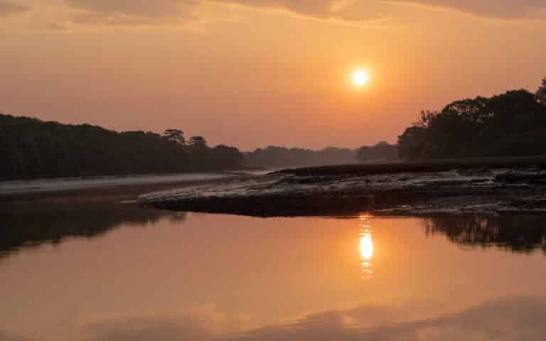A serene river scene at sunset on the Bellarine Peninsula, with the sun casting an orange glow. Trees line the riverbanks, and calm water reflects the vivid colours above—perfect for those seeking new home builds in top suburbs.