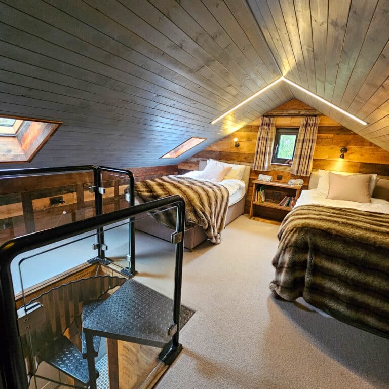 Cosy attic bedroom with sloped wooden ceiling, two single beds with fur blankets, wall lights, flat roof windows to brighten your home, a small window, and a staircase with glass balustrade. Warm, rustic decor and natural light from rooflights.