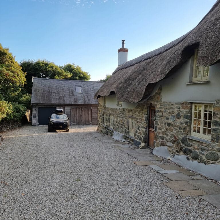 A stone cottage with a thatched roof stands beside a gravel drive, leading to a wooden garage where a black car is parked. Trees and greenery surround the property, offering great potential for veranda improvement under the clear blue sky.