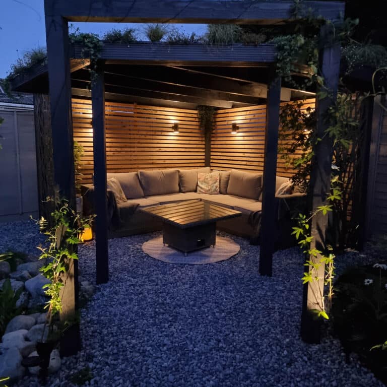 A cosy outdoor seating area under a wooden pergola at dusk, softly lit by warm lights. Inspired by modern veranda designs, it features a corner sofa with cushions and a fire pit table, all surrounded by gravel and lush greenery.